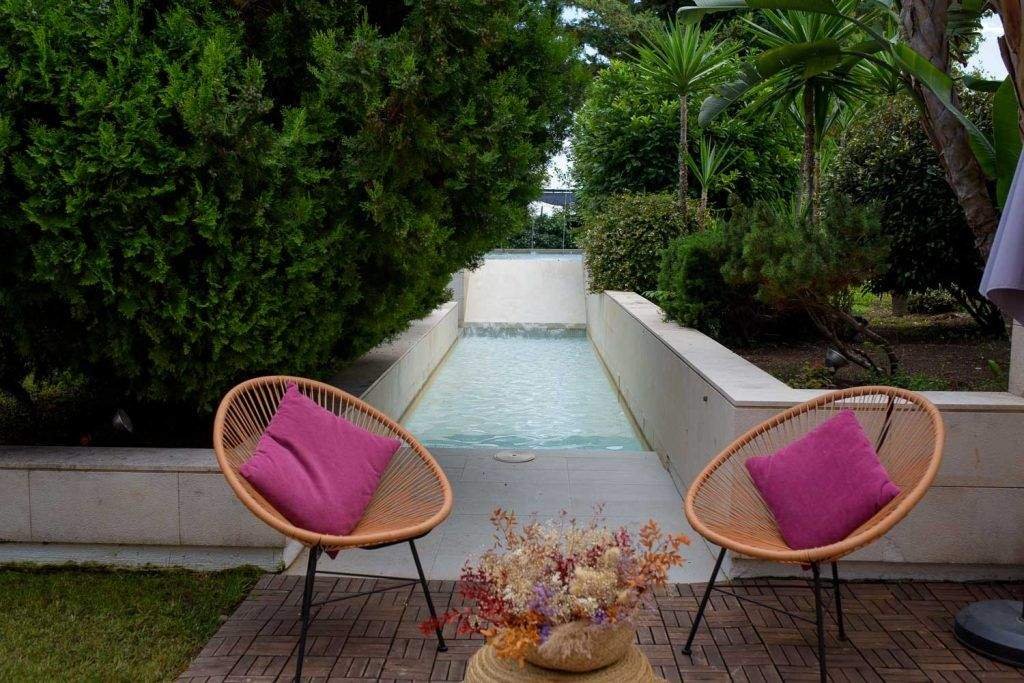 Dos sillas redondas de caña con respaldos en un jardín con piscina decorativa detrás y vegetación a los lados.